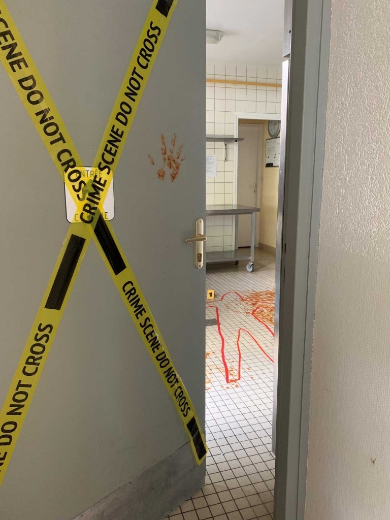 Il y a une porte avec des rubans avec marqué "Do not cross" (ne pas traverser). La porte est entrouverte et on y voit une marque de cadavre avec du sang.
